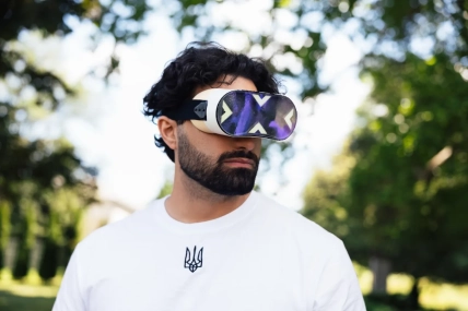 Бородатый мужчина в белой футболке с трезубцем в VR-очках на фоне деревьев