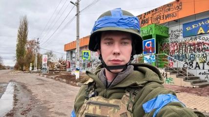 Молодий чоловік у касці та бронежилеті стоїть на вулиці біля будівлі з графіті