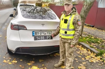 Чоловік у військовій формі та світловідбивному жилеті тримає чорний номерний знак біля задньої частини білої Tesla