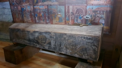 Закрита старовинна дерев’яна труна з хрестом на кришці стоїть на підставках перед розписаною стіною