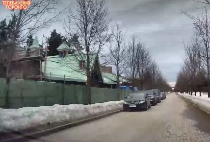 Дом с зеленой крышей за забором, вдоль заснеженной улицы стоят припаркованные автомобили