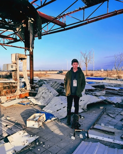 Молодий чоловік із бейджем стоїть серед уламків зруйнованої будівлі, перед ним встановлена відеокамера