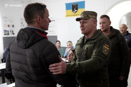 Військовий у формі розмовляє з чоловіком у куртці, позаду сидять і стоять люди