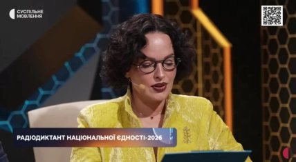 Даша Астафьева в круглых очках и желтом жакете с брошью читает текст с папки в студии