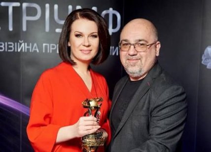 Женщина в красном платье с наградой и мужчина в очках стоят рядом на фоне баннера