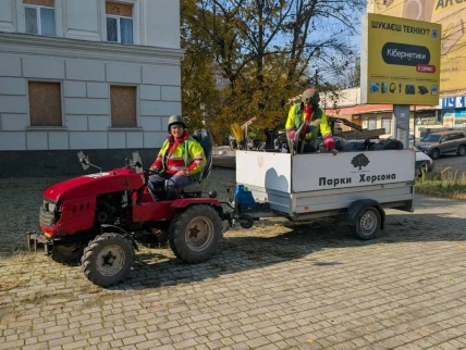 Двоє працівників у світловідбивному одязі на червоному тракторі з причепом на вулиці Херсона