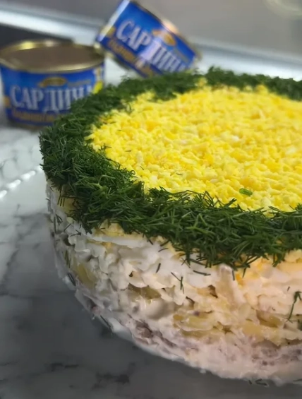 Слоеный салат с желтым верхом и зеленью по краю, две банки сардин на заднем плане