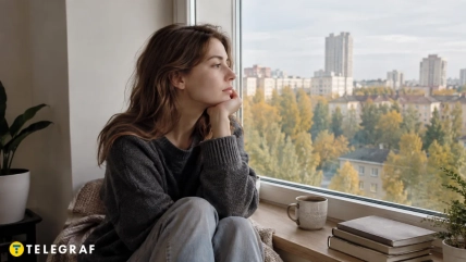 Девушка в тёмном свитере сидит у окна с чашкой и книгами