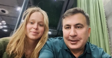 Мужчина в бирюзовой рубашке и светловолосая девушка сидят рядом перед камерой