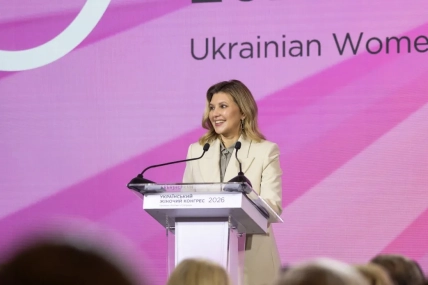 Олена Зеленська у світлому жакеті та сірій сорочці виступає за трибуною на рожевому тлі