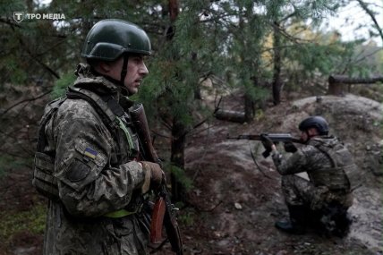 Військовий у камуфляжі та шоломі стоїть із автоматом, інший прицілюється з автомата в лісі