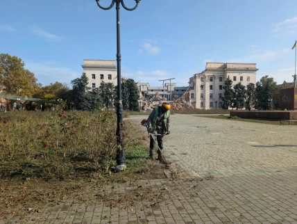 Працівник із тримером біля ліхтаря на площі; за ним – частково зруйнована будівля